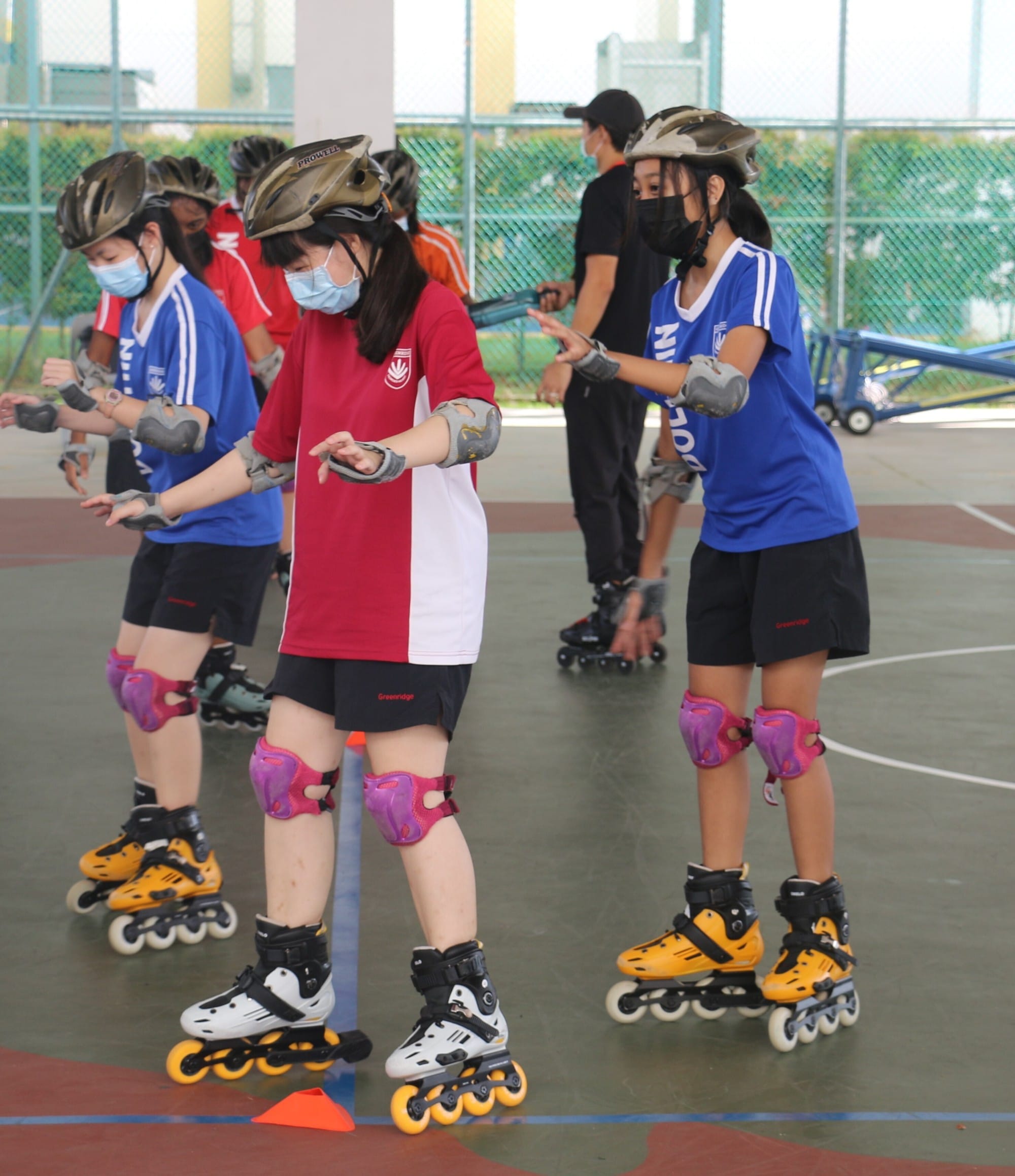 Students wearing helmets, masks, and knee pads practice inline skating on an outdoor court.